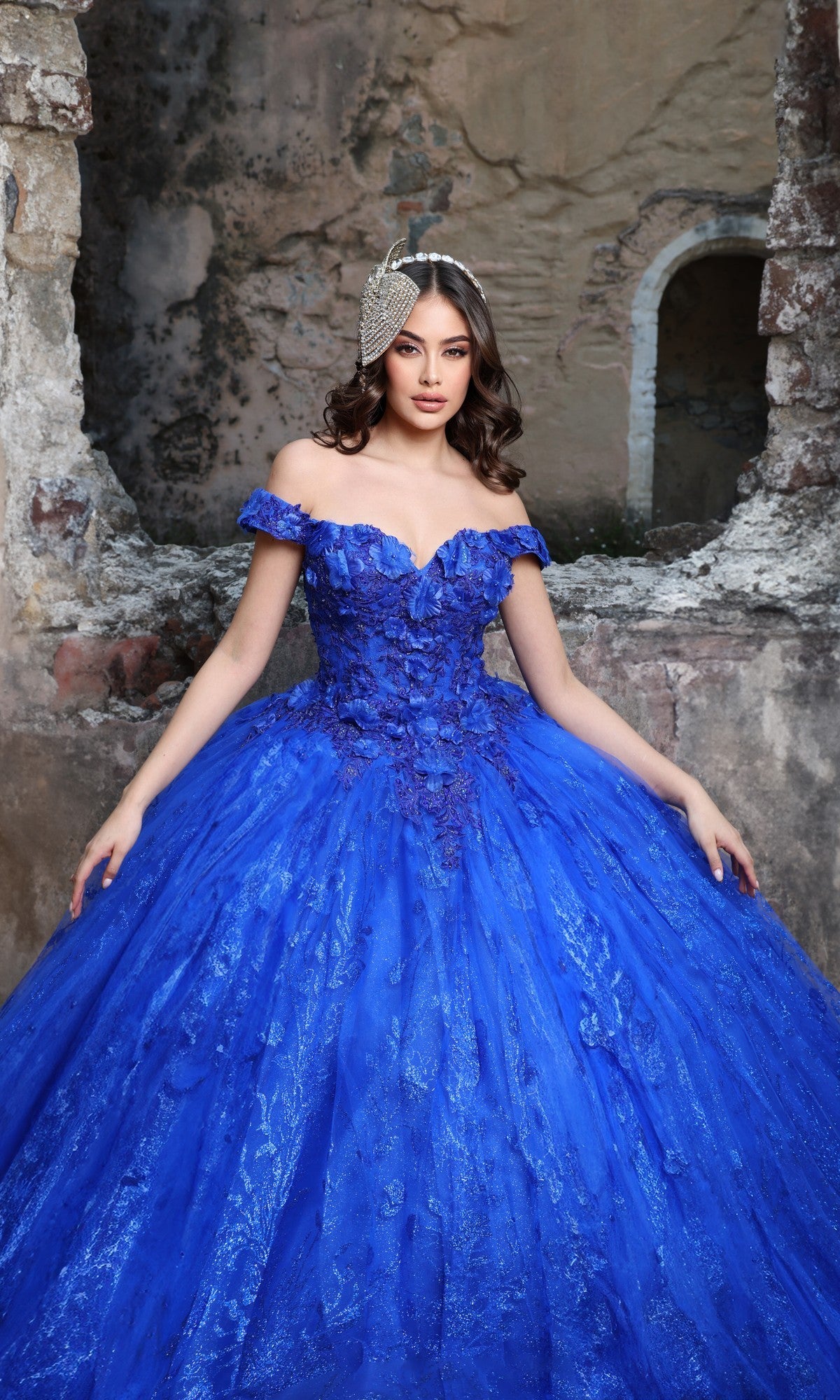 A woman wearing the Quinceanera Dress Q129 by Nina Canacci in royal stands before a weathered stone wall, adorned with a jeweled headpiece and gazing at the camera—a strikingly elegant moment.